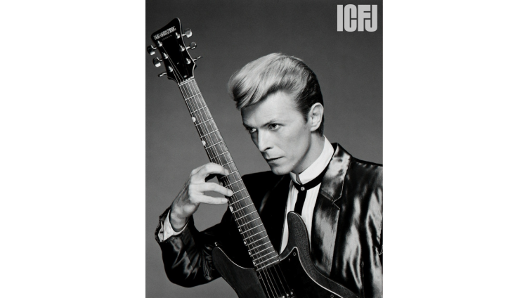 A black-and-white portrait of musician David Bowie. He’s looking to the left with a focused expression and holding his guitar upright.