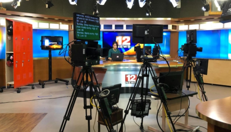 A photo of a broadcast newsroom, with teleprompters and cameras in the foreground and an anchor and anchor desk in the background.